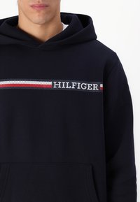 Felpa blu navy con tasca frontale, caratterizzata da un design a righe in rosso, bianco e blu, e il logo "HILFIGER" in lettere bianche.