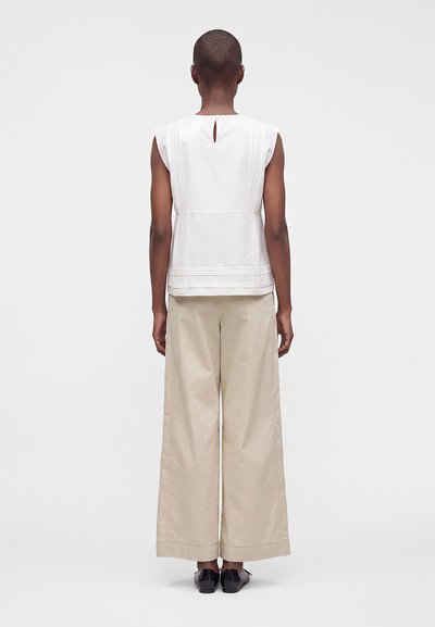 Personne aux cheveux courts portant un haut blanc sans manches et un pantalon large beige, debout, dos tourné, sur un fond uni.