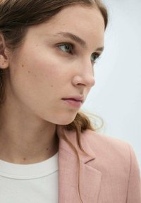 Gros plan d'une personne portant un blazer en lin rose clair sur un pull blanc. Les cheveux sont ondulés et bruns, avec un fond neutre.