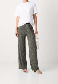 T-shirt blanc avec un logo discret, associé à un pantalon large à motifs noir et crème. Le mannequin tient un sac ceinture de couleur claire et porte des talons à lanières.