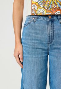 Jeans de tiro alto en azul claro, con pierna recta, que presentan un botón de cobre, costuras visibles y un ligero desgastado en la tela.