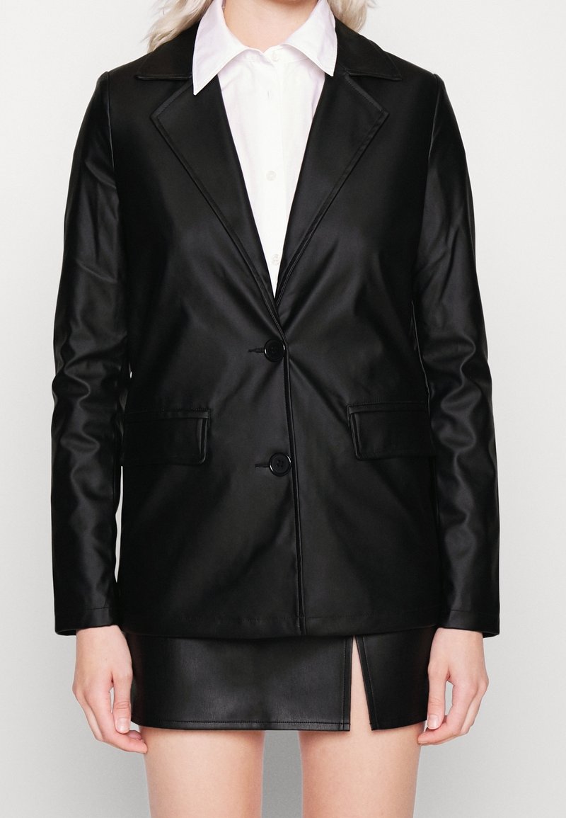 Femme portant un blazer en similicuir noir avec deux boutons et une mini-jupe assortie avec une fente devant, associée à une chemise blanche à col.