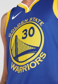 Maglia da basket dei Golden State Warriors blu e gialla con il numero 30 e il logo del ponte, indossata da una persona con un braccio tatuato.