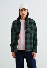 Chemise en flanelle à carreaux verts et noirs avec deux poches poitrine, boutons-pression, portée sur un T-shirt rose et un pantalon en denim foncé. Casquette blanche sur la tête.