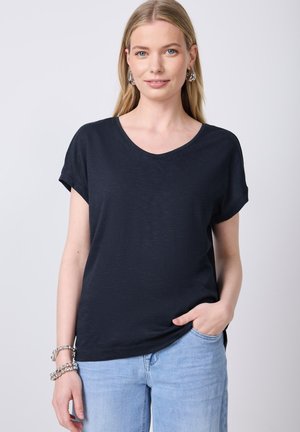 T-shirt nera a maniche corte con scollatura a V, realizzata in tessuto morbido. Il modello la indossa con jeans azzurri chiari, con una mano in tasca.