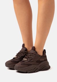Hnědé sportovní tenisky s síťovanými panely, silnými gumovými podrážkami a kovovými akcenty na tkaničkách. Design se vyznačuje zakřivenými liniemi a texturovaným povrchem.