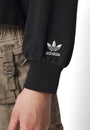 Close-up van een hand met een zwarte Adidas-trui met een wit trefoil-logo op de boord, gecombineerd met beige cargobroeken met een metalen gesp.