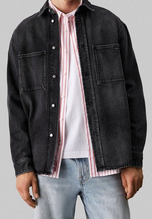 Mann trägt mehrere Kleidungsschichten, darunter eine schwarze Jeansjacke, ein weißes Hemd mit roten vertikalen Streifen und hellblaue Jeans.