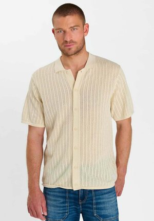 Man draagt een beige gebreid overhemd met korte mouwen, knopen en een kraag, gecombineerd met een blauwe spijkerbroek, tegen een effen witte achtergrond.