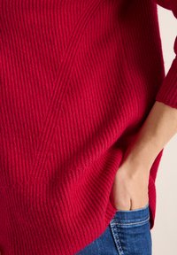 Roter, gerippter Strickpullover mit lockerer Passform, der ein diagonales Nahtdesign und lange Ärmel aufweist, kombiniert mit blauen Jeans.