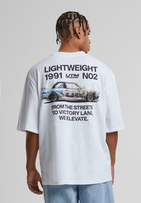 Bjela oversized majica s grafikom šarenog automobila i natpisom. Mekani pamučni materijal s kratkim rukavima i opuštenim krojem.