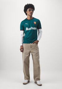 Teal Puma-tröja med svarta detaljer och vit "Auchan"-logotyp, lager på lager över en vit långärmad skjorta, ihopparat med beige byxor och vita sneakers.