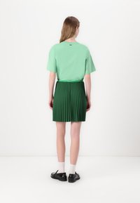 Grønn kortermet topp kombinert med en plisserte mørkegrønn skjørt, tettsittende midje, og svarte sko med hvite sokker. Enkel og strukturert design.