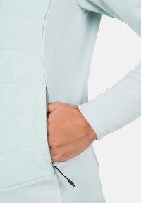 Lyseblå sportsbluse med en sidetask med lynlås, lange ærmer og teksturerede stofpaneler nær fronten. Justerbar snøre detaljer ved lommen.