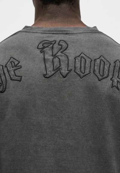 Πίσω όψη ενός ατόμου που φορά μια ξεθωριασμένη γκρι μπλούζα με μαύρο γοτθικό κείμενο κεντημένο στη μέση της πλάτης.