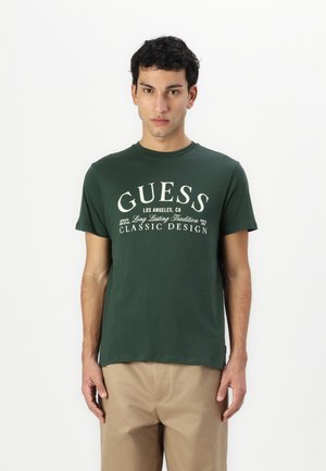 Temnozeleno bombažno majico s krožnim ovratnikom, ki ima na sprednji strani napisano belo tiskano besedilo "GUESS CLASSIC DESIGN" v krepki, ukrivljeni pisavi.
