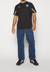 Černé sportovní tričko s modrým logem na hrudi a žlutými akcenty na rukávech, kombinované s modrými volnými džíny a černými botami.