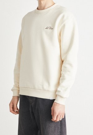 Personne portant un sweat crème avec le logo "Les Deux" sur la poitrine et un pantalon gris foncé sur un fond blanc uni.