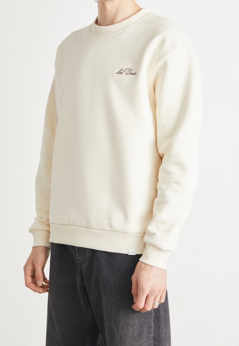 Personne portant un sweat crème avec le logo "Les Deux" sur la poitrine et un pantalon gris foncé sur un fond blanc uni.