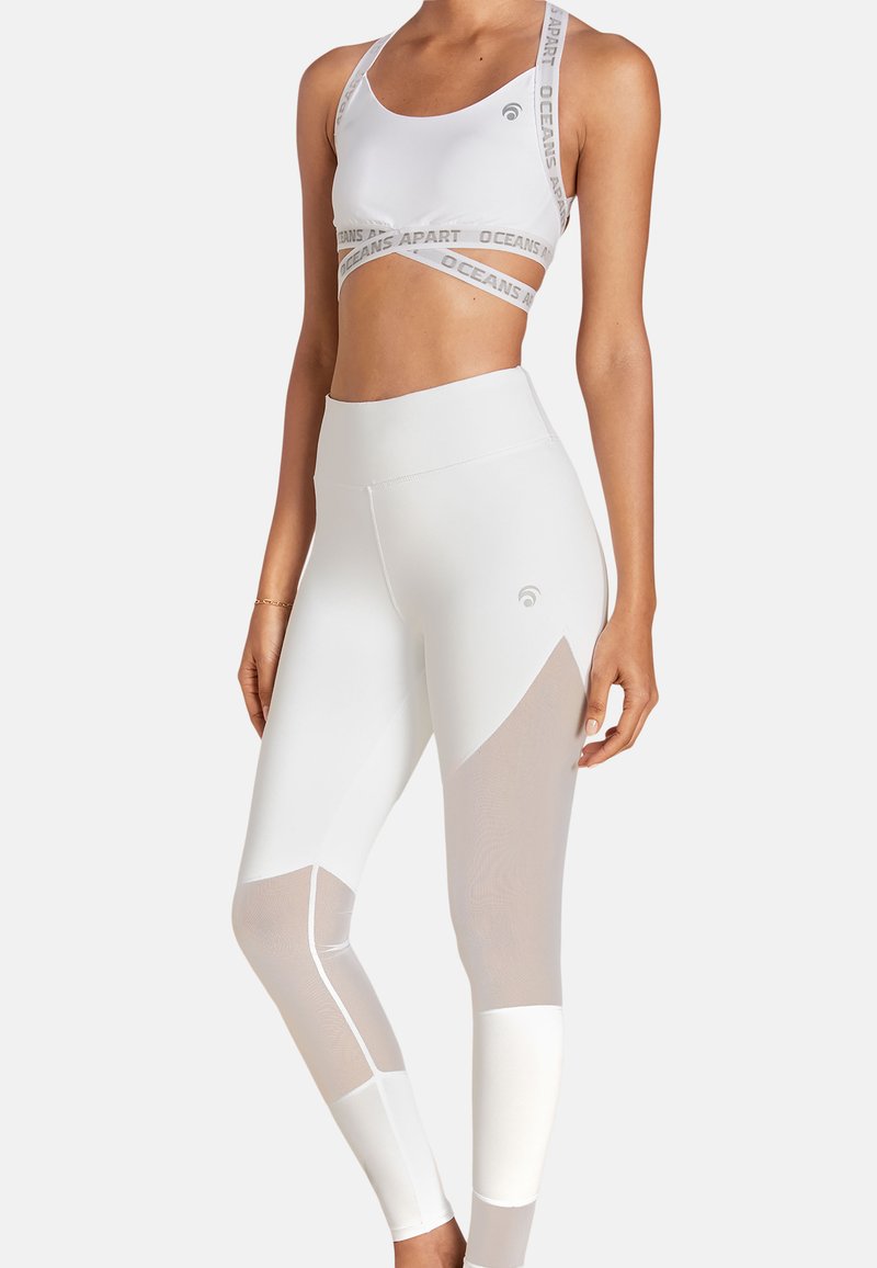 Crop top atletico bianco e leggings a vita alta con dettagli in mesh; il tessuto presenta pannelli a contrasto e branding sulla banda superiore.
