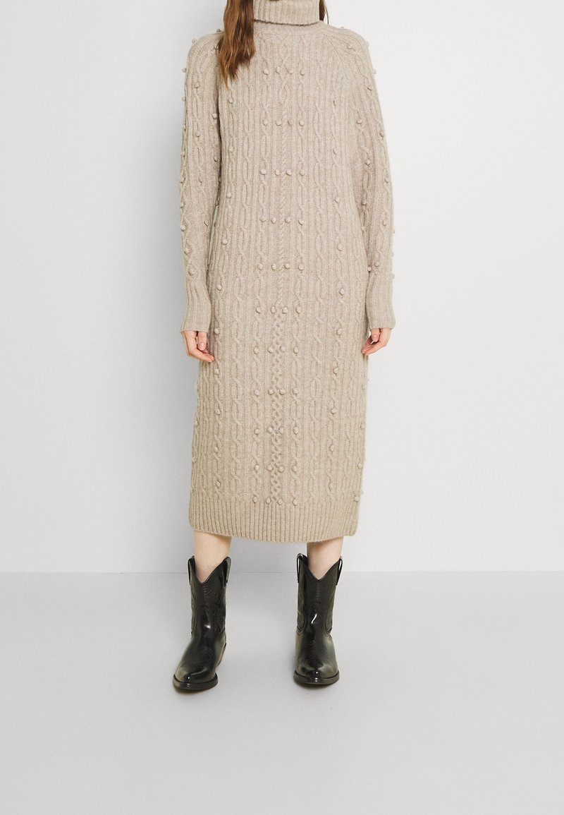 Femme portant une robe midi beige en maille torsadée col roulé avec des détails en boules et des bottes de cow-boy noires mi-mollet, debout contre un mur uni.