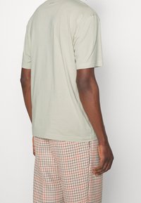 Muž v svetle šedé košili s krátkým rukávem a béžových kostkovaných kalhotách, stojící proti jednoduchému bílému pozadí, zády ke kameře.