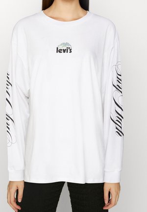 Personne portant une chemise blanche à manches longues Levi's avec un logo de montagne sur la poitrine et un texte manuscrit noir le long des deux manches, assortie à un pantalon noir.