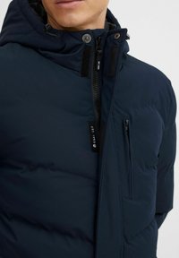 Chaqueta acolchada de color azul marino con capucha, cierre de cremallera y detalles de Velcro. Cuenta con un bolsillo lateral y una etiqueta que dice "EST. 1993". Textura suave.