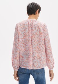 Blusa trasparente di un rosa chiaro con un motivo astratto arancione, maniche lunghe con polsini elasticizzati, abbinata a jeans blu.