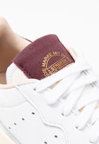 Vita lädersneaker med en vinröd mocka hälflik, guldfärgade detaljer, mesh-tunga och vita snören. Har små perforeringar längs sidorna.