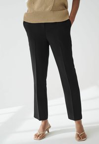 Zwarte op maat gemaakte broek van gladde stof, met een rechte snit, zijzakken en een net afgewerkte look. Gedragen met een beige gebreid topje en sandalen.
