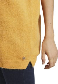 Gros plan sur la main d'une personne à côté d'un pull tricoté jaune avec un petit logo argenté près de l'ourlet, assorti à un pantalon bleu foncé.