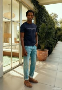 Laivastonsininen puuvillainen t-paita, valkoinen logo, vaaleansiniset farkut ja ruskeat kengät kirkkaan vihreää ulkotilaa vasten.