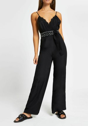 Kobieta ubrana w czarny kombinezon z szerokimi nogawkami, z koronkowym dekoltem, wzorzystym paskiem w talii, kolczykami kołami i czarnymi płaskimi sandałami ze złotymi akcentami.