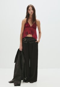 Giovane donna che indossa un top senza maniche rosso, pantaloni neri larghi con una cintura chiodata, mentre tiene una giacca di pelle nera in una mano.