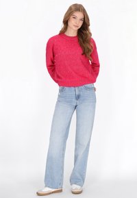 Élénk rózsaszín kötött pulóver lekerekített nyakkivágással, apró díszítésekkel. Világoskék, bőszárú farmernadrággal és fehér sportcipőkkel társítva.