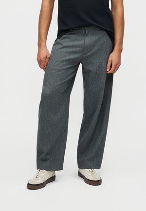 Homme portant un pantalon gris ample, des baskets blanches et une chemise noire, debout devant un fond clair uni.