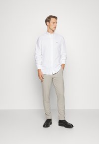 Camicia bianca con bottoni e maniche lunghe, pantaloni beige chiaro su misura e mocassini neri. La camicia presenta un piccolo logo sul petto.