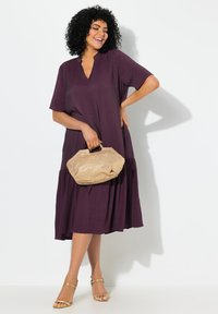 Keselamatan hingga lutut, gaun longgar berwarna plum kaya dengan lengan pendek dan leher V dalam. Dipadukan dengan tas tangan tekstur berwarna beige dan sandal emas.