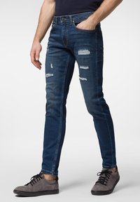 Smala denimjeans i mörkblått med slitna lagningar. Har fem fickor, knäppning med knapp och kombineras med grå sneakers.
