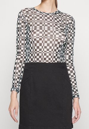 Mujer vestida con una blusa de manga larga a cuadros en blanco y negro, metida dentro de una falda negra lisa, con un fondo liso.