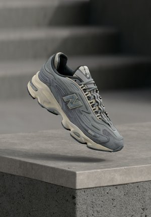 Baskets New Balance 1000 grises avec semelle, lacets et logo couleur crème, flottant au-dessus d'un rebord en béton avec des escaliers flous en arrière-plan.