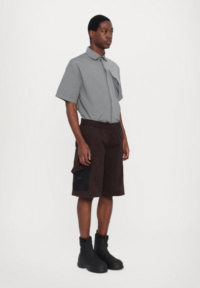 Chemise grise à manches courtes avec détail de poche, associée à un short marron foncé avec une poche latérale et des accents noirs, portée avec des bottes noires.