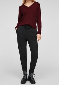 Frau trägt einen burgunderfarbenen, langärmligen Pullover mit V-Ausschnitt, schwarze Jogginghosen mit Kordelzug und schwarze Schn ürstiefel vor einfachem Hintergrund.