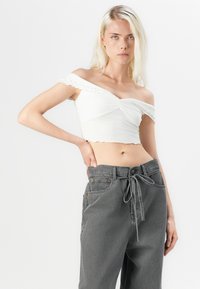 Bijela crop majica s otvorenim ramenima, s rubom od volana i twist dizajnom sprijeda, u kombinaciji s visokim sivim hlačama koje imaju remen za vezanje.