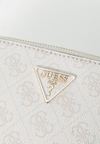 Portafoglio in tessuto bianco e testurizzato con un logo triangolare dorato "GUESS" con punto di domanda, cuciture decorative e una cerniera argentata.