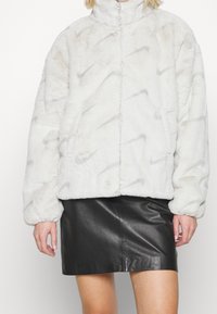 Bílá faux kožešinová bunda s vysokým límcem, zdobená jemným vzorem loga Nike. Nositá přes černou koženou sukni.