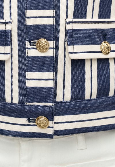 Ύφασμα με ναυτικό μπλε και λευκές ρίγες με χρυσά διακοσμητικά κουμπιά και ραμμένες λεπτομέρειες τσέπης σε ένδυμα, πάνω από λευκό παντελόνι.