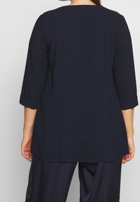 Blouse bleu foncé avec manches trois-quarts, encolure dégagée et détails de poignets retroussés. Tissu lisse avec une coupe décontractée et une ourlet droit.