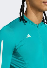 Turkusowa sportowa bluzka na zamek z fakturowanej tkaniny. Posiada białe logo Adidas oraz paski na rękawach, podkreślając dopasowany krój.